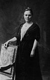 A posed formal black and white photograph of a woman standing behind a chair upon which she is resting her hand