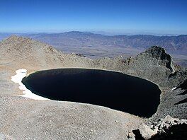 Tulainyo Lake with nearly black water, rimmed with snow, seen from above looking out at the lake and Owens Valley beyond the ridge to its east