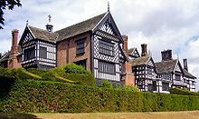 The exterior of a large house. There are several chimneys, leaded windows and wings. In the foreground are two rows of hedges.