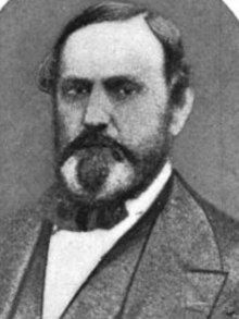 A man with receding black hair, sideburns, a mustache, and a goatee wearing a black jacket, vest, and bowtie, and a white shirt