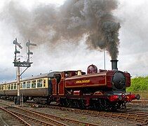 A pannier tank locomotive is pulling two cream and brown passenger carriages. The locomotive is mainly painted maroon, but there is black and yellow lining on the side of the pannier tank, the cab, the bunker, the toolbox, and the splashers. The words "LONDON TRANSPORT" are shown in yellow on the side of the pannier tank. The chimney and front of the smokebox are black. The front buffer beam is red.