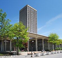 A single modern-style tower is surrounded by a lower open-air pavilion with trees accenting the area.