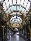 A wide shopping precinct, seen from within the interior, with glass roofs, detailed artwork to the cornices, and shop fronts to either side