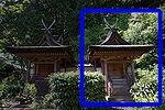 A small wooden building next to another identical building with gabled roof, a stair on the gable side covered by an extended roof. The roof ridge has forked finials.