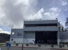 A fairly square-shaped, tall, gray building with glass entrance doors surrounded by large glass panes. Text on the side read NAVAL UNDERSEA MUSEUM.