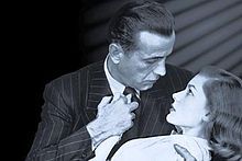 Black-and-white image of a man and a woman, seen from mid-chest up, their faces in profile, gazing into each other's eyes. He embraces her in a dip with his right arm and holds her right hand to his chest with his left hand. He wears a pin-striped suit and a dark tie. She wears a white top. On the left, the background is black; on the right, it is lighter, with a series of diagonal shadows descending from the upper corner.