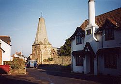 Street scene showing stone church with truncated spire, On the right is a white painted building.