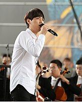 Min Kyung-hoon singing into a microphone with an orchestra in the background