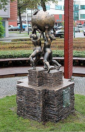 Four figures stand in a circle holding up the symbol of Wikipedia, which consists of a sphere made of jigsaw puzzle pieces, with few pieces missing, symbolizing that the work of the encyclopedia will never be finished