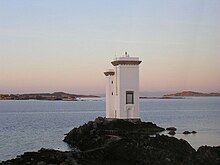 A white building consisting of a single white tower four stories high and an adjacent tower appearing to be a single storey lower, sits on a rock overlooking the sea. The sun is not visible but is evidently low in the sky as pinkish light suffuses the landcape. In the distance there are a myriad low rocky islets.