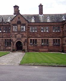 Part of a two-storey stone building seen directly from the front. A paved path leads to a doorway in a slightly protruding bay; over the doorway is an inscribed plaque, and above that is a statue in a niche.