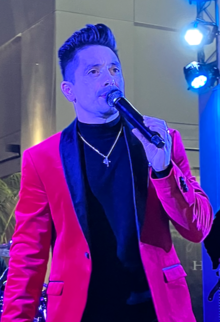 A man with short dark hair, wearing a black shirt and red jacket with black lapels, holding a microphone