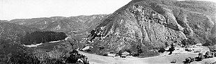 Black and white panoramic view of a canyon, a group of trees is to the left and several residential buildings are at lower right.