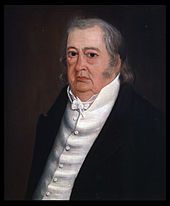 A man with long, gray, thinning hair wearing a white button-up shirt and a black jacket. He is facing left.