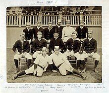 Two rows of men, one standing behind one seated, of men wearing old-style white baseball uniforms and striped pillbox caps