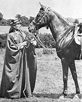 A black-and-white photograph of a European woman dressed in Bedouin robes and head covering, standing in front of a dark horse equipped with a bridle and saddle.