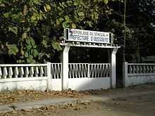 A white gate with a sign reading "Republique du Senegal: Prefecture d'Oussouye"