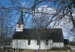 A small white church with white brick walls, brown windows and a black roof. It is standing near trees having no leaves and a graveyard. As the picture was taken, it was slightly cloudy.