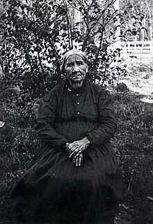 An elderly woman wearing a patterned dress sitting in front of a bush with her hands in her lap.