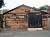 A low plain building in red sandstone with a gently sloping gabled roof and a door to the right with flaking black paint and a notice reading "Bridewell" in Gothic script