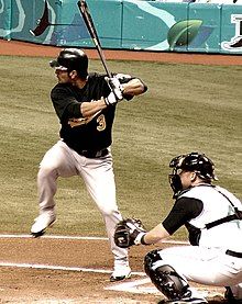 A man in a dark baseball jersey, batting helmet, and white pants takes a left-handed baseball swing while another man kneels wearing catcher's gear behind him.