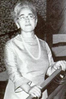 An older woman in harsh light, her grey hair in a bouffant updo, wearing a gown with long flaired sleeves and trapunto trim details; also wearing a strand of beads and glasses