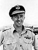 Outdoor half portrait of grinning man in light-coloured uniform with pilot's wings on left pocket, wearing peaked cap