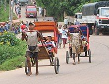 Two Malagasy men pull children in rickshaws. Both modern vehicles and more rickshaws can be seen in the background.