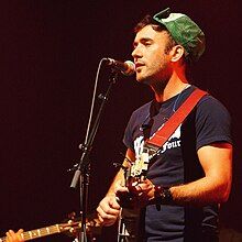 A young Caucasian man with short, dark hair plays a banjo.