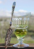 A reservoir glass filled with absinthe next to an absinthe spoon.