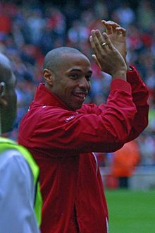 Freddie Ljunberg, wearing gloves and a redcurrant football shirt applauds the crowd. A stand full with people and man wearing a football shirt are visible in the background.