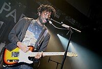 A male vocalist plays electric guitar in front of a mic stand. The stage is lit by white lights, and its background is mostly black.