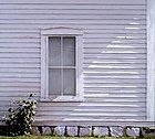 old white clapboard house, purplish hue case by shade, with window in center reflecting sky, stone base surrounded by weeds carefully rendered