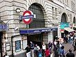 A street filled with people in front of a light grey building that has variously coloured signs protruding from it stating a variety of different things