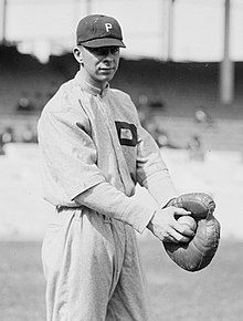 A black-and-white image of a man wearing a white old-style baseball uniform and holding a baseball with his right hand in the catcher's mitt on his left hand