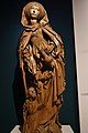 A wooded sculpture of St. Elizabeth. She has on a 15th-century style dress and wears a cloak which falls to mid shin. She holds a pitcher in her left hand and at her right foot a small depiction of a man kneels and gazes up at her.