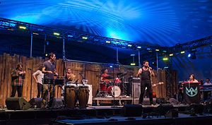 Eight men performing on a stage. From left to right; two horns, timbales, shekere, drums, vocals, bass (obscured), keyboards.