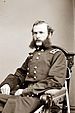 A white man with wavy hair and extremely bushy sideburns connected to a mustache, sitting in a chair. He is wearing a long double-breasted military coat with a rectangular patch over each shoulder.