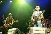 Three men on a stage, two in the forefront standing behind microphone stands and playing guitars and the third in the background sitting behind a drum set.