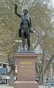 bronze statue of a man in 18th-century costume, standing with right arm raised, set on a red stone pedestal in an urban setting with trees behind it, with inscription in gold-coloured lettering beginning "BURKE 1774-1780"