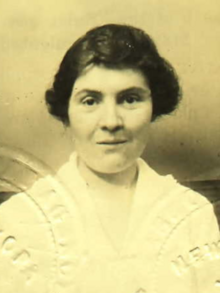 A white woman with dark hair, wearing a white top