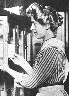 A white woman, standing, in profile, with blond hair in a bouffant style, wearing a striped dress and an apron