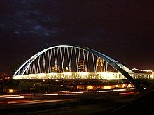 A large arched suspension bridge passes over a busy highway. The nighttime skyline is seen through the cable supports.