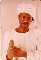 shown is Al-Amin Kabashi, a Sudanese adult man dressed in a white jalabiya, a traditional Sudanese garment, with a white turban neatly wrapped around their head. Kabashi has a calm and thoughtful expression, and their right arm is amputated just below the elbow. Kabashi gently holds the amputated arm with their left hand.