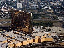 A tall, reflective, bronze-colored hotel tower sits atop a smaller structure, next to a parking garage. A highway and city streets are visible in the background.