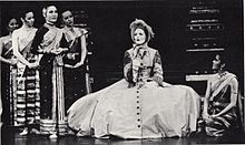 Black-and-white photo of a stage scene. A Caucasian woman wearing a bonnet sits on a chair at center, with her huge Victorian hoop skirt spread out around her, singing. Several women in elegant silky Asian clothing surround her. One at right (Tuptim) kneels watching her, as the others stand in a clump at left.