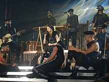 Madonna sitting onstage singing, surrounded by her dancers in a circle.