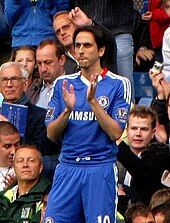 A photograph of a white man wearing blue association football attire while clapping his hands. Behind the player, several spectators can be seen looking at him.