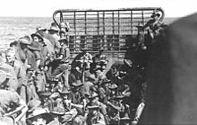 About forty troops wearing slouch hats and carrying rifles packed into a landing craft travelling across open water, viewed from the stern towards the bow ramp.