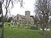 Stone building with square tower. In the foreground are gravestones and trees.